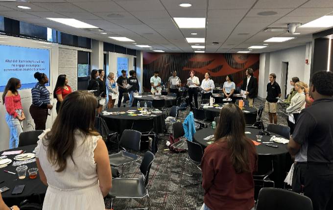 A group of 25 students standing in a circle listening to one person reflect on their experience.