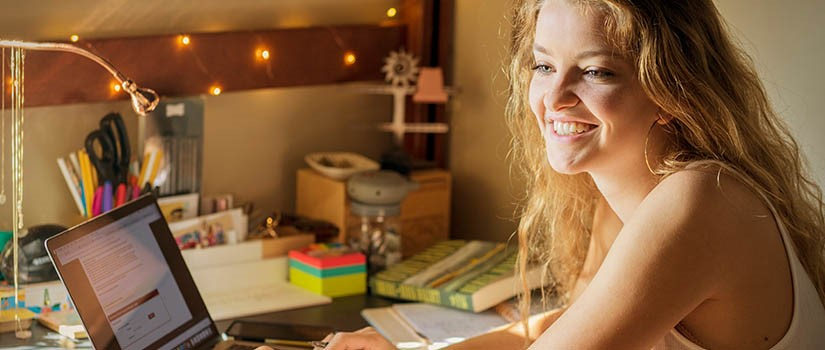 A student sits at a desk in front of her computer. She is smiling at the screen. 