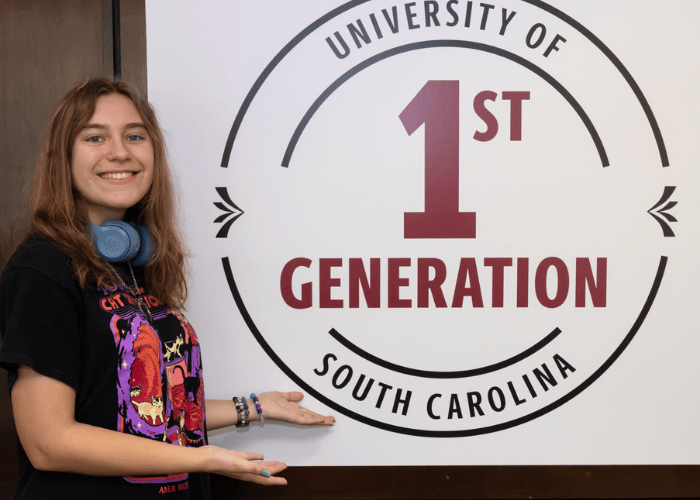 First-Gen student standing next to the USC First-Gen stamp logo (sign)