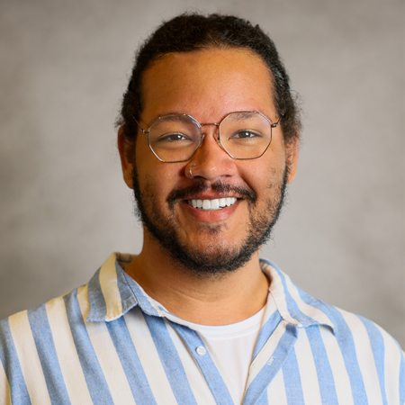 Professional headshot of a smiling person with dark hair pulled back wearing round metal eyeglasses, a neatly trimmed beard and mustache, and a blue-and-white vertically striped button-down shirt over a light-colored T-shirt. The person is facing the camera and is wearing a small nose ring. The photo is set against a soft gray studio background with warm, even lighting, creating a friendly, approachable, and professional appearance.