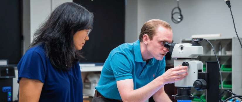 photo of professor looking on while a student looks into a microscope