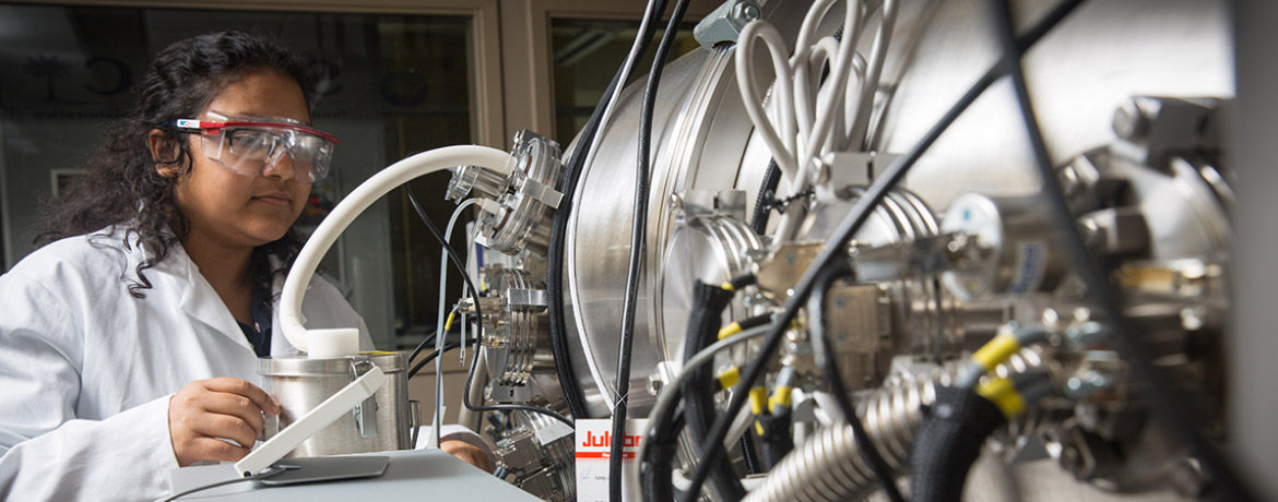 Female student in white lab coat and safety glasses works on a large piece of stainless steel equipment in a lab