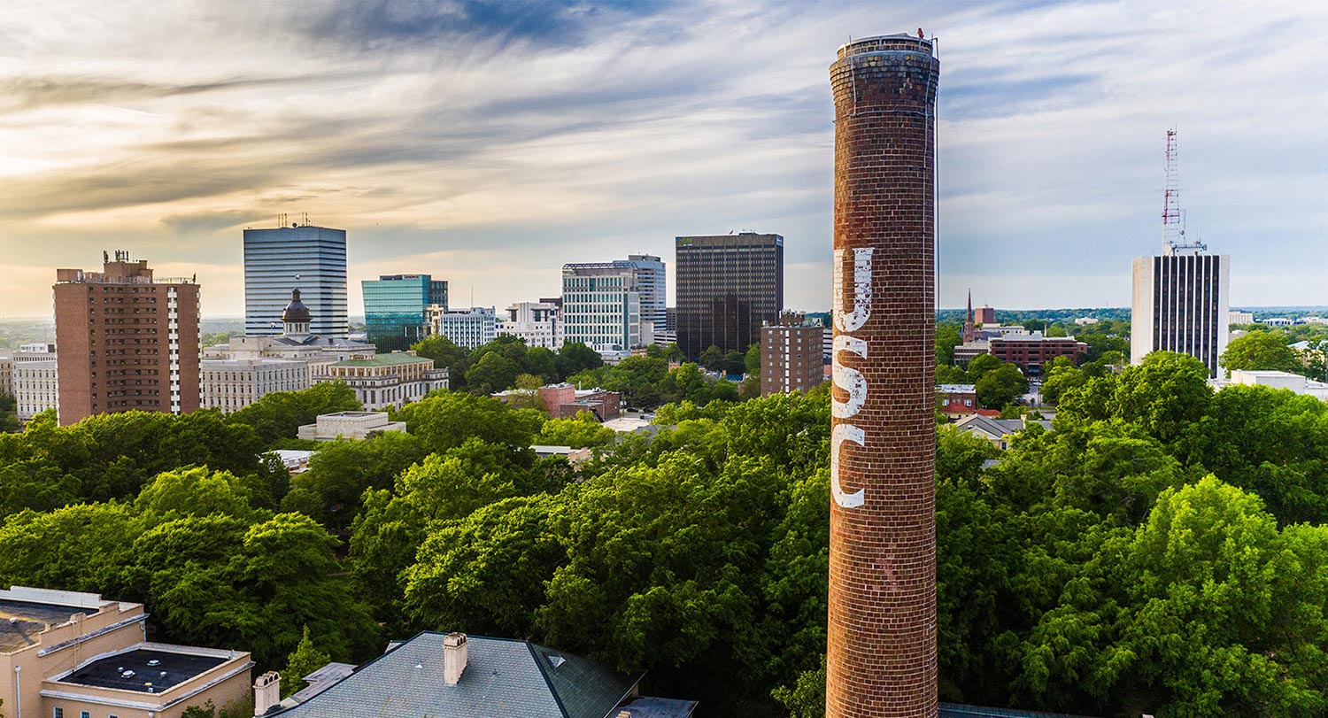 An aerial view of the University of South Carolina campus with tall buildings in the background and a brick smokestack with the letters USC printed vertically.