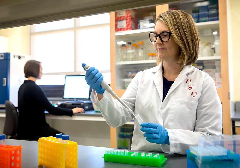 Student in a lab coat holding a pipet at a table with colorful trays. 