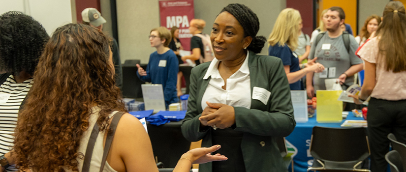 A faculty or staff member talks with a student at an academic engagement fair, surrounded by tables with program materials and other attendees.
