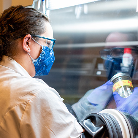 a woman wears a lab coat and a mask while working with nuclear lab equipment