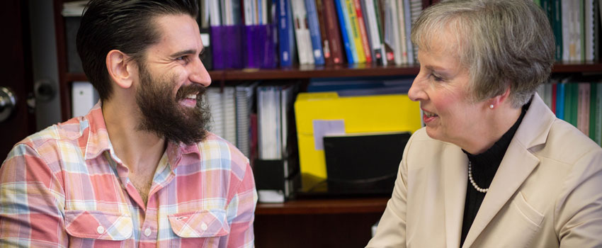 A student and professor confer at a desk in front a a stack of books.