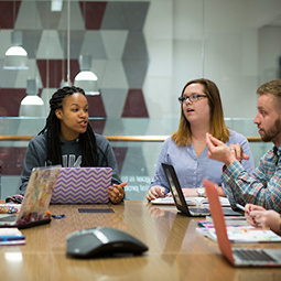 Three students, two females and a male, of mixed ethnicity having a discussion around around a table with laptops in front of them.