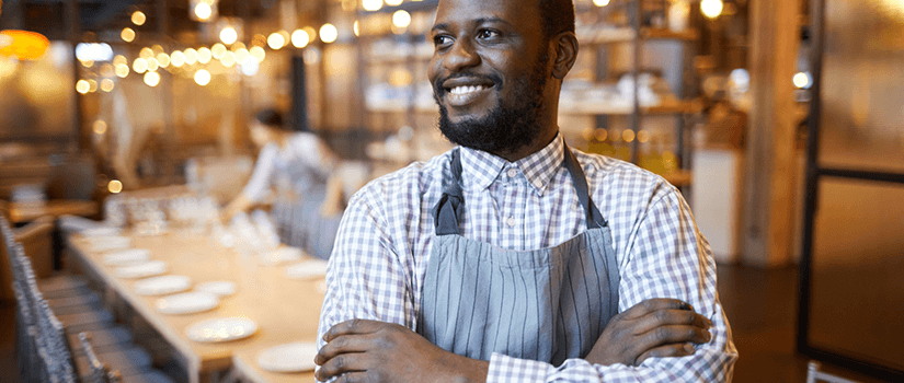 Smiling restaurant manager stands in foreground of well-lit, modern and atmospheric restaurant. 