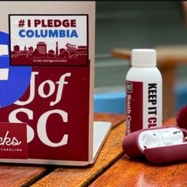 A laptop on a wooden table that has a sticker with the #ipledgecolumbia logo. There is also a hand sanitizer bottle and airpods on the table.