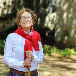 woman wearing glasses in white shirt with red sweater tied around shoulders and holding a walking stick with foliage in the background