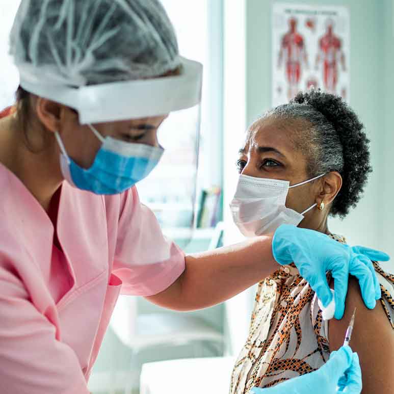 A woman dressed in protective medical gear gives an arm injection to a female patient