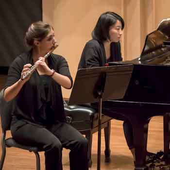 A female musician plays the flute next to a female musician playing piano