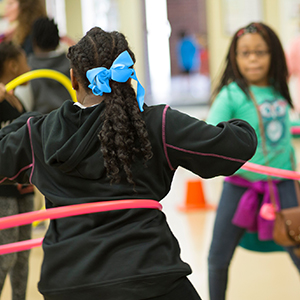 School girl hula-hooping in a gym setting with other classmates.