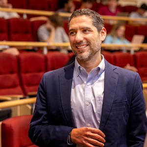 a man stands in an auditorium and speaks to two men with red theater chairs and people in the background