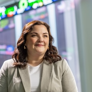 USC alumna Stephanie Kyle smiles warmly while standing in front of a financial data display board showing stock tickers